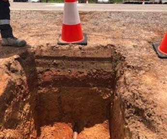 Underground utility work beneath Stuart Highway, showing a freshly dug trench with exposed piping and safety precautions in place.